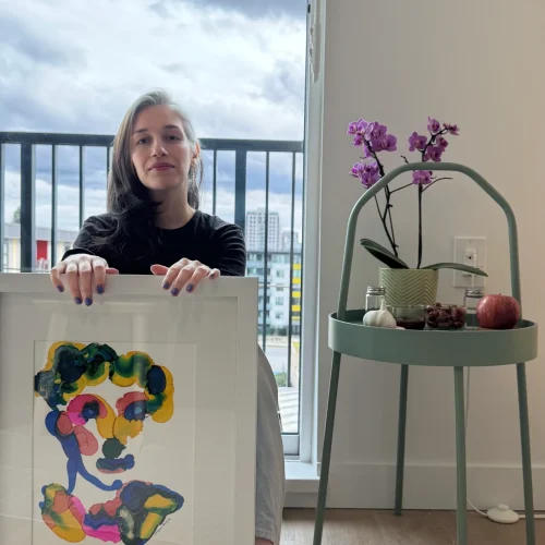 Visual artist Moojan Azar holding her framed alcohol ink portrait artwork while seated indoors beside a purple orchid and city balcony.