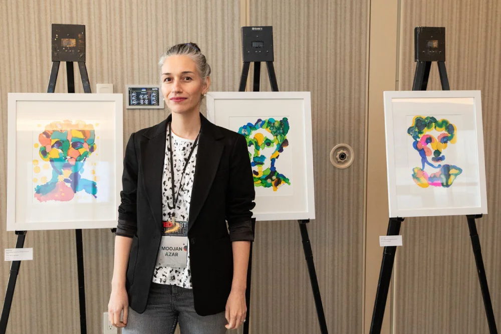 Moojan Azar standing in front of three colorful abstract portraits at her TEDxBTT exhibition at the Paradox Hotel, Vancouver.